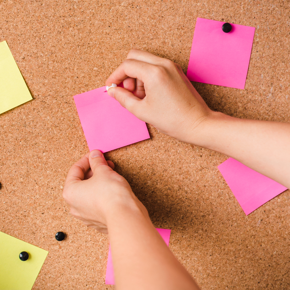 close-up of a person fixing pink adhesive note with thumbtack on corkboard